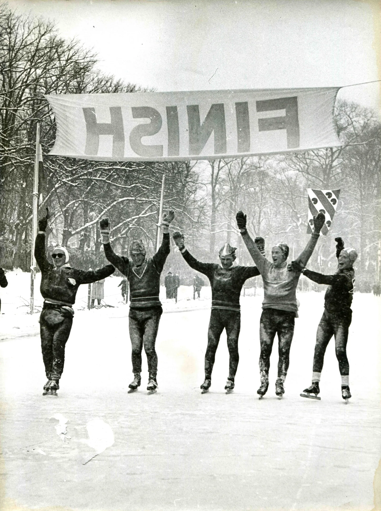 Vijf juichende schaatsers steken hun armen omhoog onder een FINISH-spandoek aan het einde van de Elfstedentocht van 1956