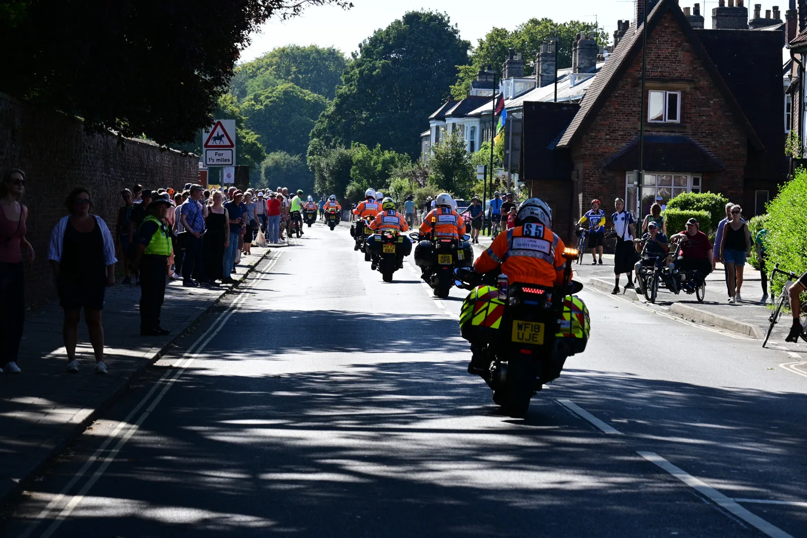 Groep motorrijders in colonne op een doorgaande weg tussen toeschouwers en huizen