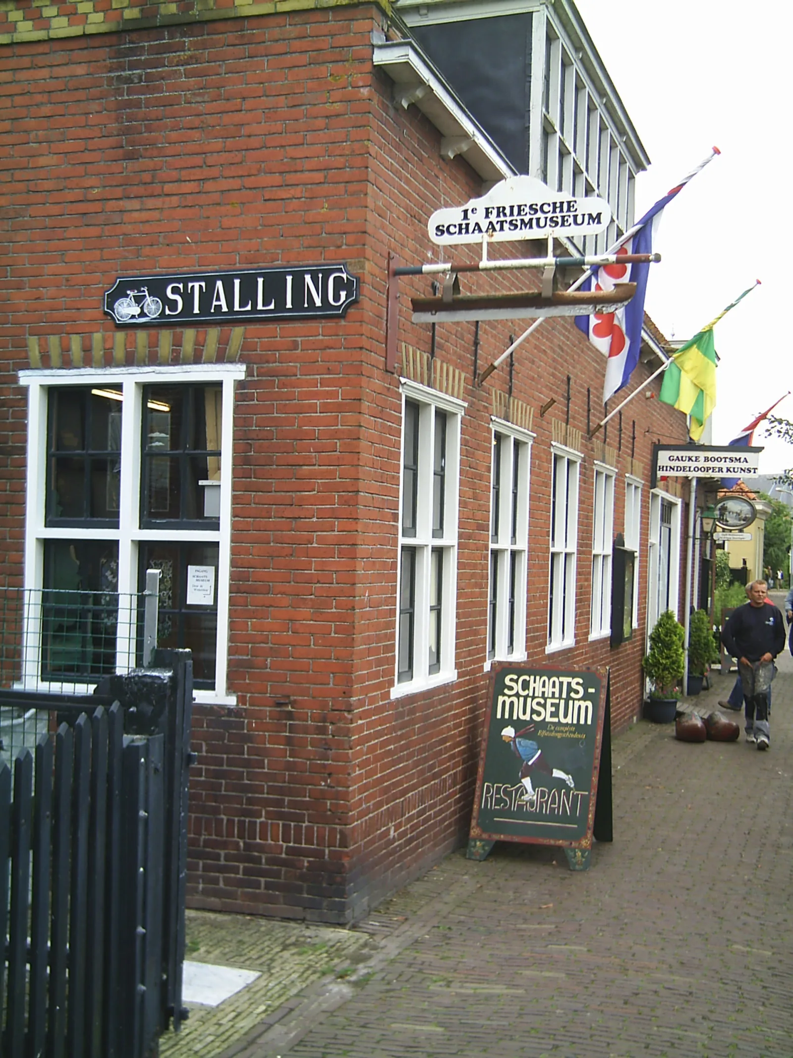 Gevel van het Eerste Friese Schaatsmuseum in Hindeloopen met bord, Friese vlag en wandschilderingen aan de straatkant