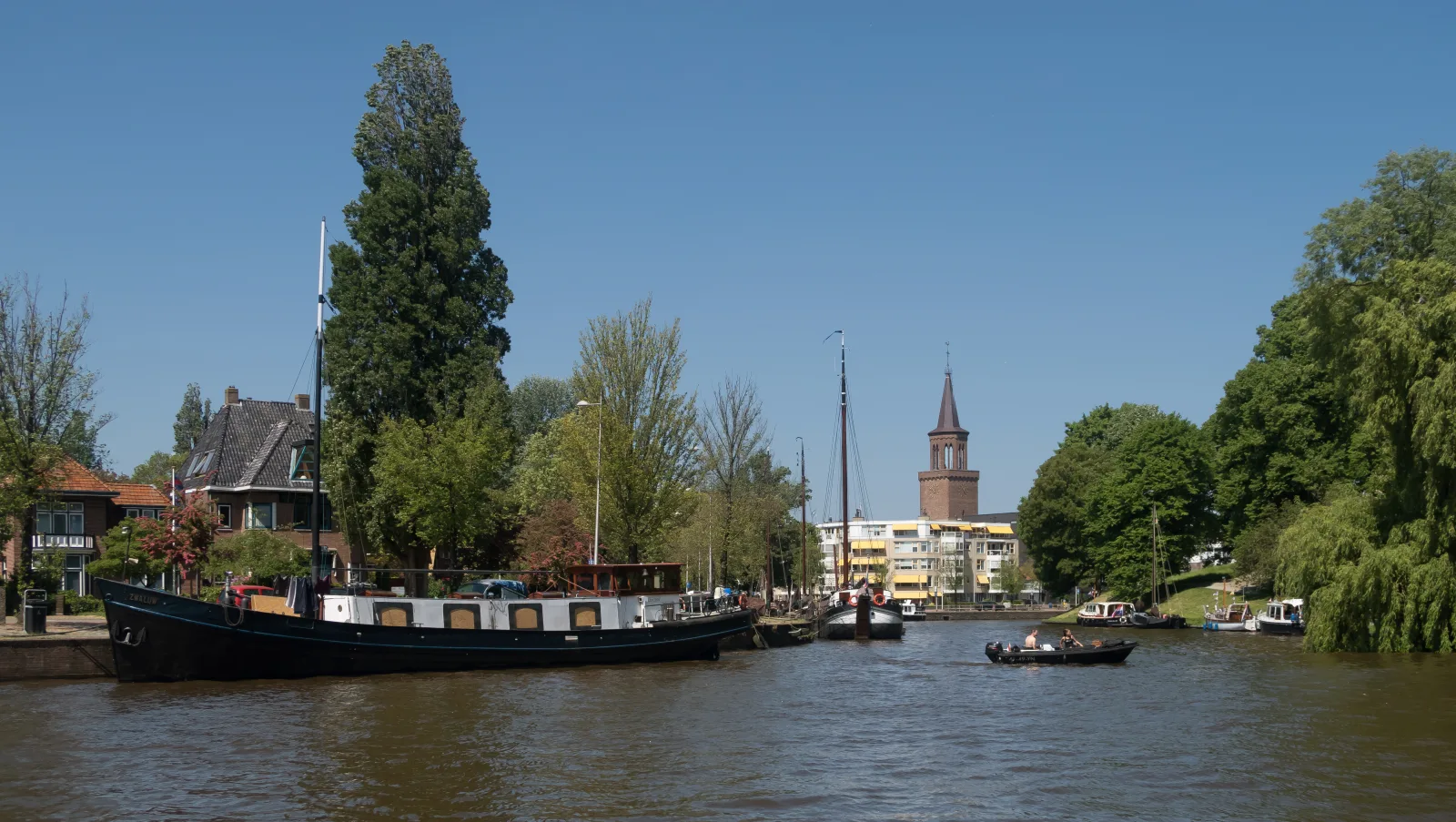 Panorama van de Wester Stadsgracht in Leeuwarden met afgemeerde schepen, bomen en de Oldehove-toren in de verte